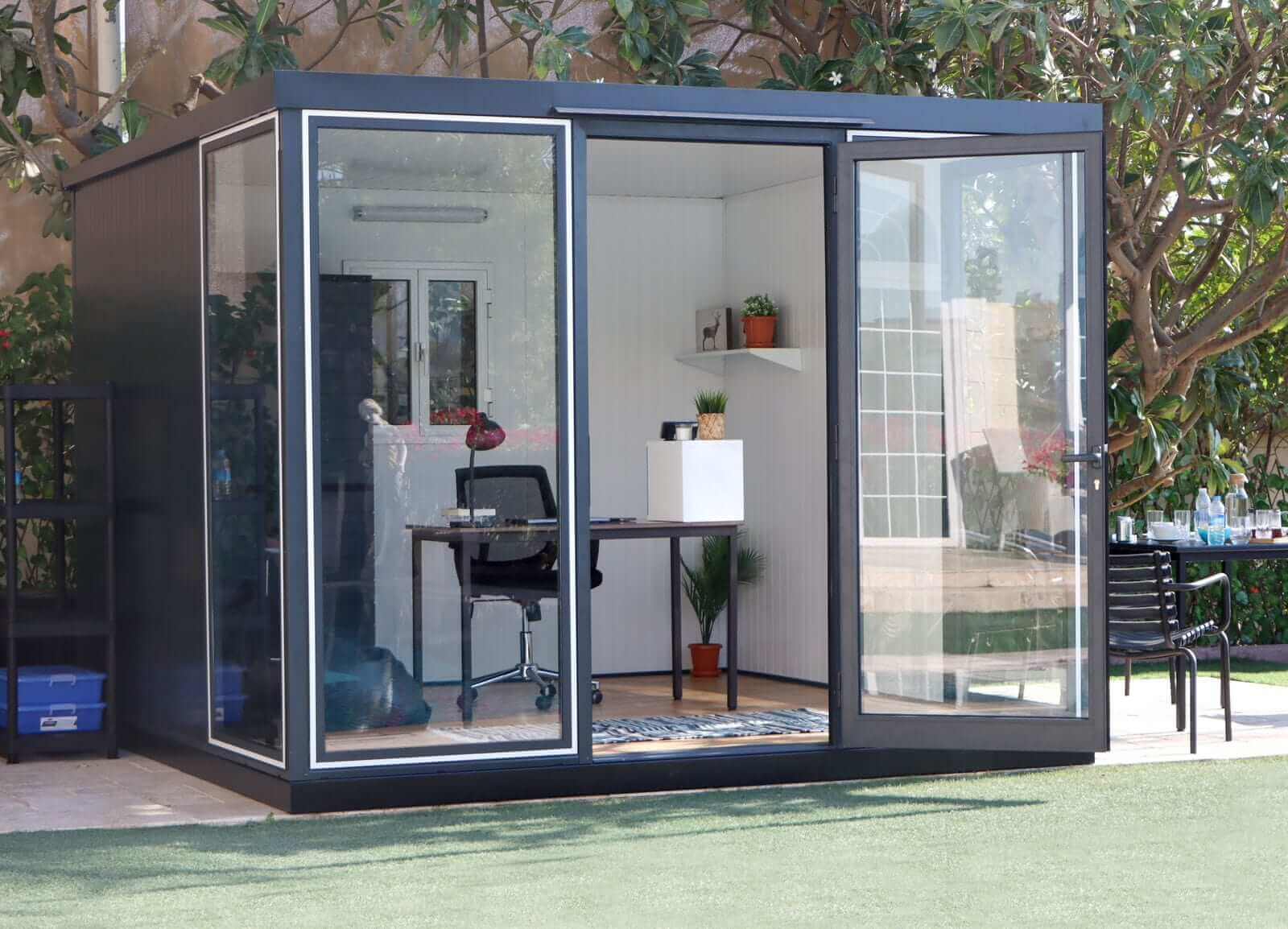 Duramax 10x10 Glass Garden Room 32001 with interior desk and chairs, surrounded by greenery.