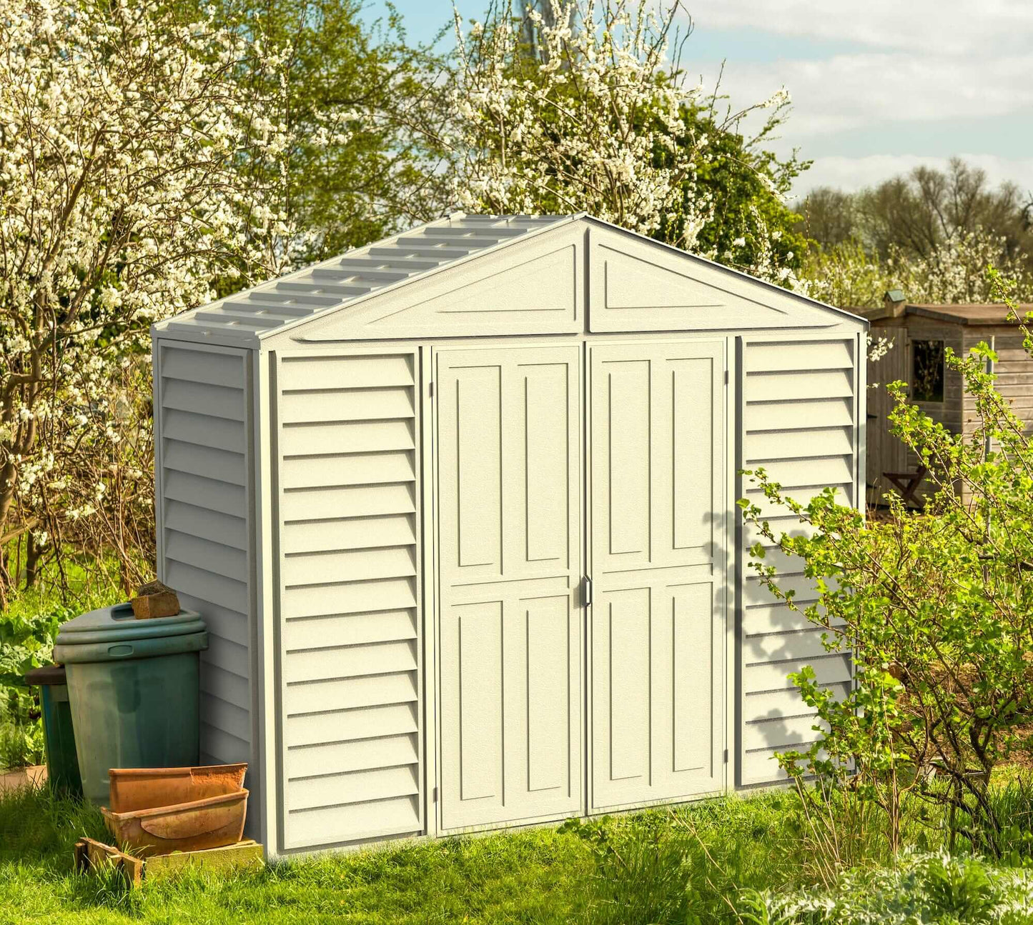 Duramax Vinyl Shed 10x3 SidePro with foundation in a garden setting, surrounded by greenery and spring flowers.