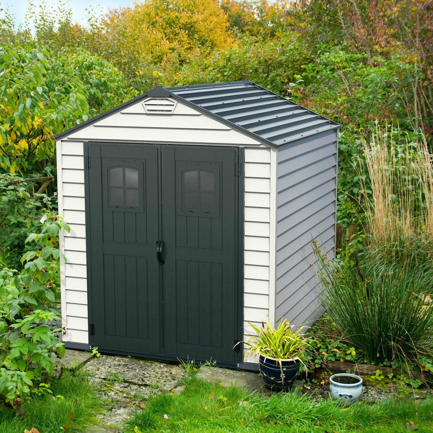 Duramax Vinyl Shed 7x7 StoreMax Plus in a garden with greenery around it.