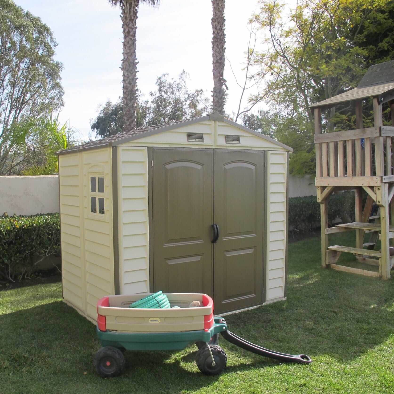 Duramax 8x6 vinyl shed in backyard with green doors, surrounded by greenery and play structure.