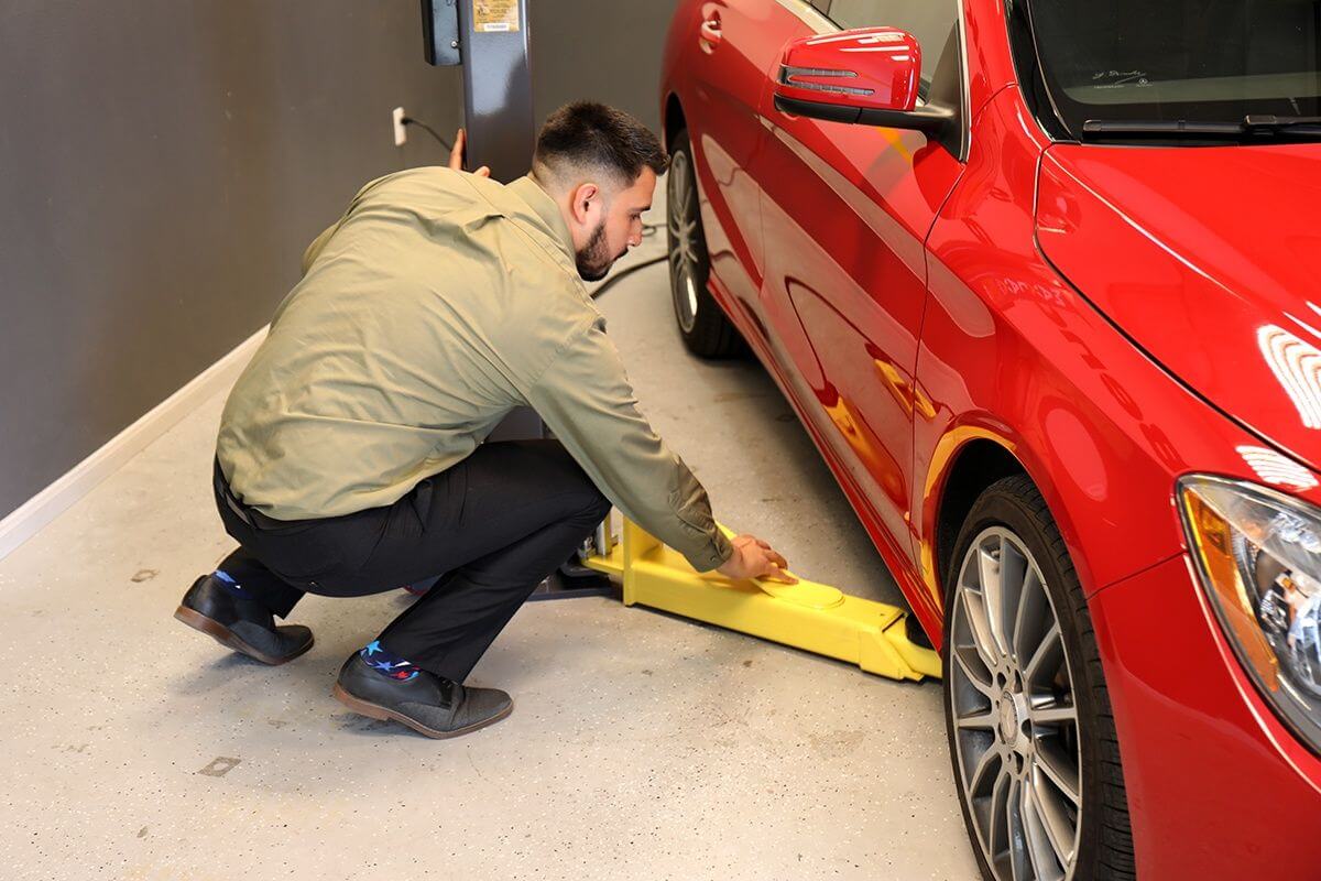 A technician using a yellow jack to lift a red car in a garage, showcasing MaxJax M7K's portability and safety features.